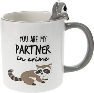 white coffee mug with text "you are my partner in crime" and an image of a raccoon painted below the text. Another 3D ceramic raccoon appears to climb out of the mug